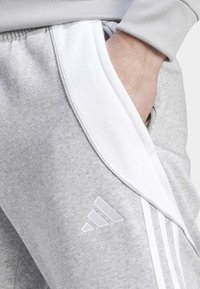 Un pantalon de survêtement gris présente une texture douce, des panneaux latéraux blancs et trois rayures blanches. Il comprend un logo brodé sur la cuisse.