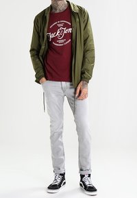 Olivgrön lätt jacka över en vinröd t-shirt med grafik, i kombination med ljusgrå jeans och svarta högklackade sneakers.