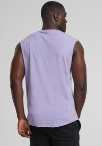 T-shirt sans manches lavande, en coton, coupe décontractée, encolure ronde, texture lisse, vue arrière avec ourlet arrondi. Pantalon noir visible.