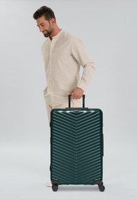 Valigia rigida di colore verde scuro con motivo a zig-zag, superficie testurizzata, maniglia superiore, maniglia estensibile e quattro ruote per la mobilità.