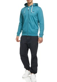 Haut à capuche zippé turquoise avec capuche et cordons blancs, associé à un pantalon foncé et des baskets blanches. Présente un logo sur la poitrine.