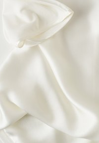 Blusa de seda en color blanco roto, con una textura suave y un diseño drapeado. Realzada con costuras delicadas a lo largo del escote y las mangas.