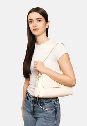 Giovane donna con lunghi capelli scuri che indossa una t-shirt bianca e jeans, tenendo una borsa a tracolla color crema con chiusura a scatto.