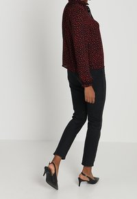 Blusa de estampado rojo y negro con cuello alto, mangas largas y jeans negros de corte ajustado. Tacones negros con tiras en el tobillo completan el conjunto.