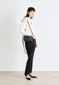 Borsa a tracolla nera con finitura texturizzata, hardware dorato e tracolla regolabile. Indossata con un hoodie crema e pantaloni neri con spaccature laterali.