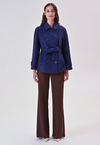 Cappotto doppiopetto blu navy con cintura, grandi bottoni e maniche larghe, abbinato a pantaloni marroni a vita alta e scarpe puntate color nude.