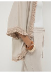 Beige fringed kimono met een zachte textuur, gecombineerd met lichtbeige broeken. Geaccentueerd met een houten armband en een doorzichtige ronde armband.