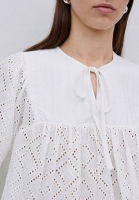 Blusa bianca con scollo rotondo e dettaglio a nastrino, caratterizzata da intricati motivi a diamante in broderie anglaise e vita arricciata.