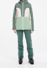 Ensemble veste de ski et pantalon en vert et beige. La veste est dotée d'une fermeture éclair devant, d'une capuche et de deux poches sur la poitrine. Le pantalon est ajusté avec des ourlets renforcés.