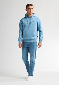 Sudadera con capucha de color azul claro con cordones, bolsillo tipo canguro y logo negro; combinada con jeans azul claro y zapatillas blancas.