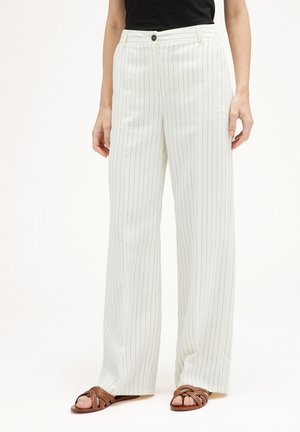 Witte gestreepte wijde broek met knoopsluiting en riemlussen, gemaakt van een lichte stof, gecombineerd met bruine sandaaltjes met banden.