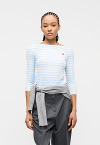 Νεαρή γυναίκα με πλεξούδες, που φοράει μπλε ανοιχτού χρώματος πουκάμισο με λευκές ρίγες μακρυμάνικο και μικρό κέντημα φράουλας, γκρι πουλόβερ δεμένο στη μέση και γκρι παντελόνι.