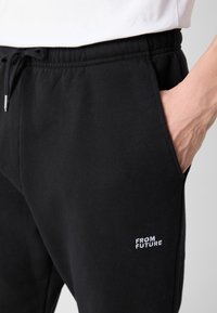 Svart joggebukser med elastisk midjebånd, sidelommer og subtil "FROM FUTURE" hvit broderi på venstre lår. Mykt materiale.