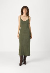 Olive krāsas, bez piedurknēm maxi kleita ar iekļautu kakla izgriezumu, izgatavota no mīksta auduma; kombinēta ar brūnām kovboju zābakiem. Gluda tekstūra un pieguļošs siluets.