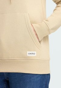 Felpa beige con tasca a marsupio e polsini a coste. Presenta un'etichetta con "oxmo" sull'orlo, abbinata a jeans in denim blu.