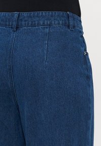 Pogled od blizu na pas i gornji deo bedara plavih traperica, prikazujući petlje za remen, šavove i ivicu bočne džepa.