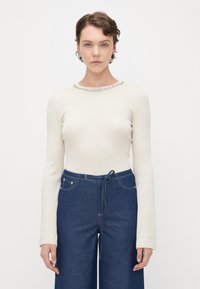Ribbed cream long-sleeve top with a round neckline embellished with metal studs, paired with high-waisted dark blue denim pants.