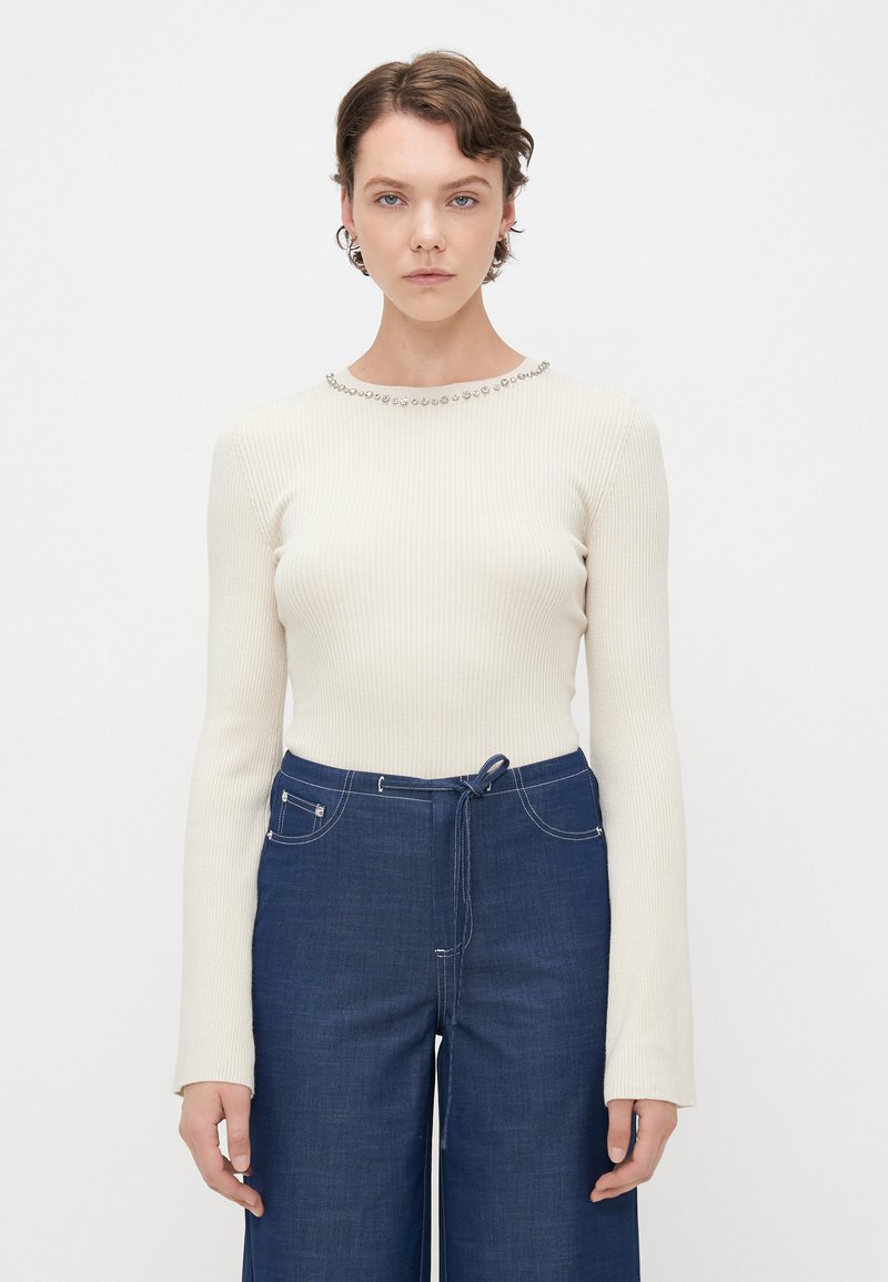 Ribbed cream long-sleeve top with a round neckline embellished with metal studs, paired with high-waisted dark blue denim pants.