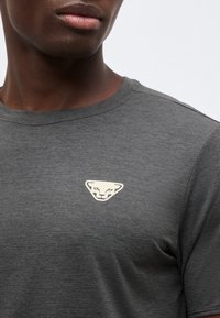 Κοντινό πλάνο ενός ατόμου που φοράει σκούρο γκρι μπλουζάκι με στρογγυλή λαιμόκοψη και μικρό λευκό στυλιζαρισμένο λογότυπο πάνθηρα στο στήθος.