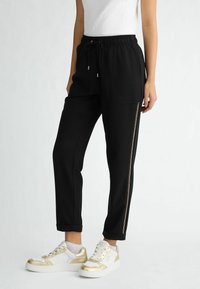 Czarne spodnie dresowe z ściągaczem w talii, bocznymi kieszeniami i zamkiem błyskawicznym jako ozdobą na nogawce. Połączone z białymi sneakersami ze złotymi akcentami.