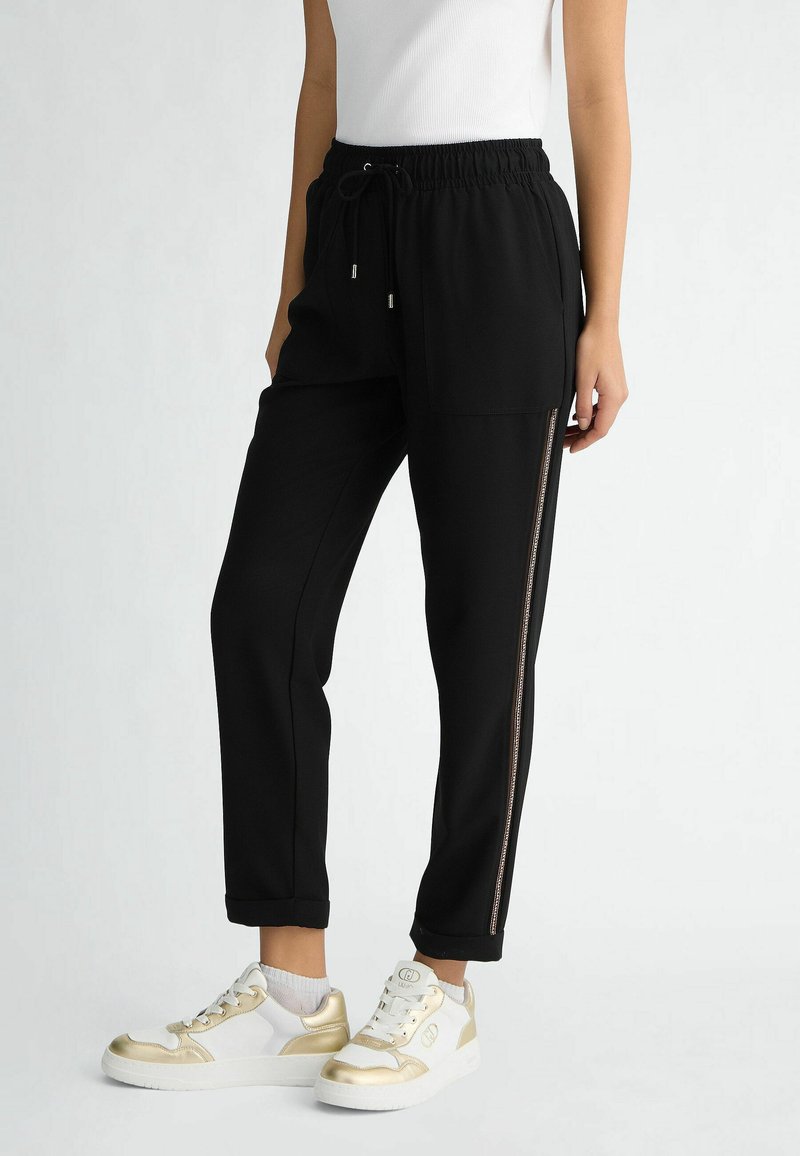 Czarne spodnie dresowe z ściągaczem w talii, bocznymi kieszeniami i zamkiem błyskawicznym jako ozdobą na nogawce. Połączone z białymi sneakersami ze złotymi akcentami.