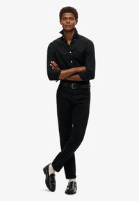 Camisa negra de botones con cuello doblado, combinada con jeans negros, un cinturón negro y mocasines negros pulidos; el fondo neutro resalta el enfoque.