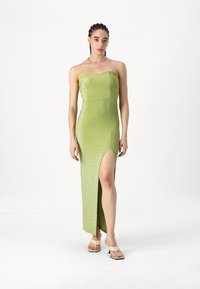 Vestido verde sem alças feito de um tecido brilhante, com uma fenda até à coxa de um lado. Usado com sandálias bege.