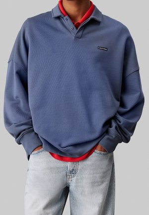 Homme portant un sweat-shirt bleu ample de Calvin Klein sur une chemise rouge, avec un jean délavé, les mains enfoncées dans les poches avant.
