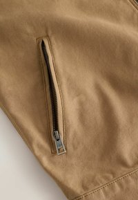 Gros plan sur une fermeture éclair métallique partiellement ouverte sur un tissu texturé beige, montrant les coutures détaillées autour de la couture de la fermeture éclair.