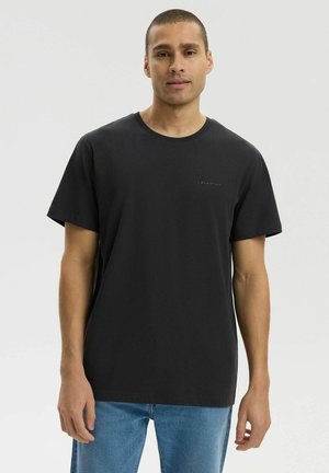 Młody mężczyzna ubrany w prostą czarną koszulkę z krótkim rękawem i niebieskie dżinsy, stojący na jednolitym jasnym tle.