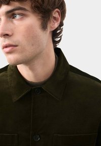 Oliwkowa kurtka z korduroyu z kołnierzykiem w kształcie punktu, dwoma przednimi kieszeniami i czarnymi guzikami. Chropowata tkanina jest dobrze widoczna, co zwiększa jej atrakcyjność wizualną.