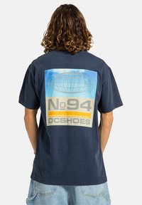 Námořnické bavlněné tričko s velkým potiskem na zádech, který zobrazuje globus, text "No94" a "DCSHOES" v žlutých a šedých akcentů.