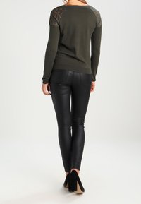 Olivgrüner Pullover mit Spitzenakzenten an den Schultern, kombiniert mit schwarzen, eng anliegenden Lederimitathosen und schwarzen High-Heels.