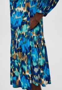 Blau- und grün gemustertes Kleid mit ausgestelltem Saum, langen Ärmeln und einer Seitentasche. Hergestellt aus leichtem, strukturiertem Stoff.