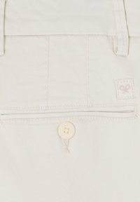 Tela de algodón beige con un bolsillo trasero con cierre de botón y un pequeño logo bordado en la esquina. Diseño simple y limpio.