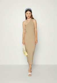 Vestido midi bege, ajustado ao corpo, com um ombro. Tecido liso e elástico. A modelo segura uma bolsa amarelo claro e usa uma faixa azul na cabeça.