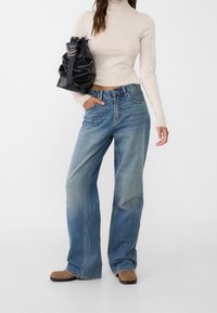 Lichtbeige coltrui in combinatie met blauwe wijde jeans met een hoge taille, subtiele vervaging, en een zwarte handtas met een geplooid ontwerp.