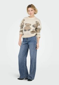 Sweter z wzorem misia w kolorze beżowym, zestawiony z luźnymi dżinsami w kolorze niebieskim. Sweter posiada prążkowane mankiety i dekolt.