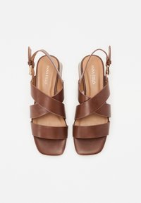 Brūni ādas sandales ar slingback dizainu, ar krusteniskām siksnām un kvadrātveida purngaliem. Iekļauta diskrēta zeltaini tonēta sprādze.