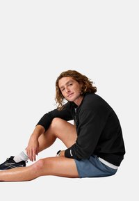Felpa nera, pantaloni corti azzurro chiaro, scarpe sportive nere e calze bianche. Pelle con lentiggini e capelli ricci. Posizione seduta su uno sfondo neutro.