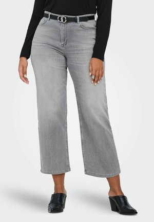 Moteris dėvi pilkus aukštos juosmens plačius sijoninius trumpintus džinsus su juodu diržu, juodą ilgomis rankovėmis palaidinę, sidabrinius žiedus ir juodus aukštakulnius mule batus.