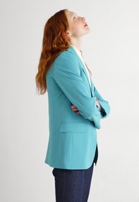 Blazer turchese con vestibilità sartoriale, un bottone e due tasche laterali. Abbinato a pantaloni scuri, mostrato su un modello di profilo.