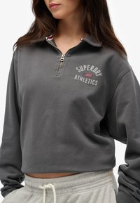 Sweatshirt zippé gris avec col rond et texte imprimé "SUPERDRY ATHLETICS". Texture douce avec une coupe courte et des accents côtelés.