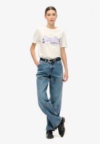 Hvid t-shirt med lilla grafisk tekst, parret med løstsiddende blå jeans sikret med et sort bælte. Sorte sko fuldender outfittet.