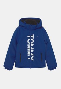 Blauwe waterdichte jas met capuchon, ritssluiting aan de voorkant en grote witte "TOMMY" tekst. Inclusief zwarte ritszakken aan de zijkanten.