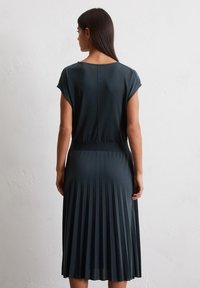Abito plissettato verde scuro con maniche corte, vestibilità rilassata e rifiniture a costine in vita e all'orlo, che mostrano una texture verticale.