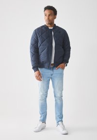 Mann som står iført en marineblå quiltet jakke, lyseblå jeans, hvite joggesko og et klokke, og ser svakt til siden.