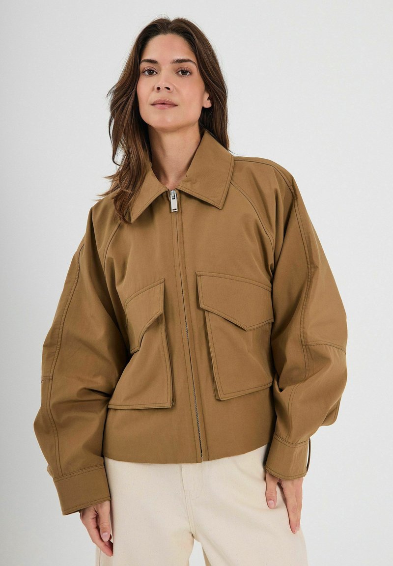 Pruun ülekaalus jakk, millel on terav krae, esine tõmblukk ja kaks suurt klapiga taskut. Sile kangas koos silmapaistvate õmblusdetailidega.