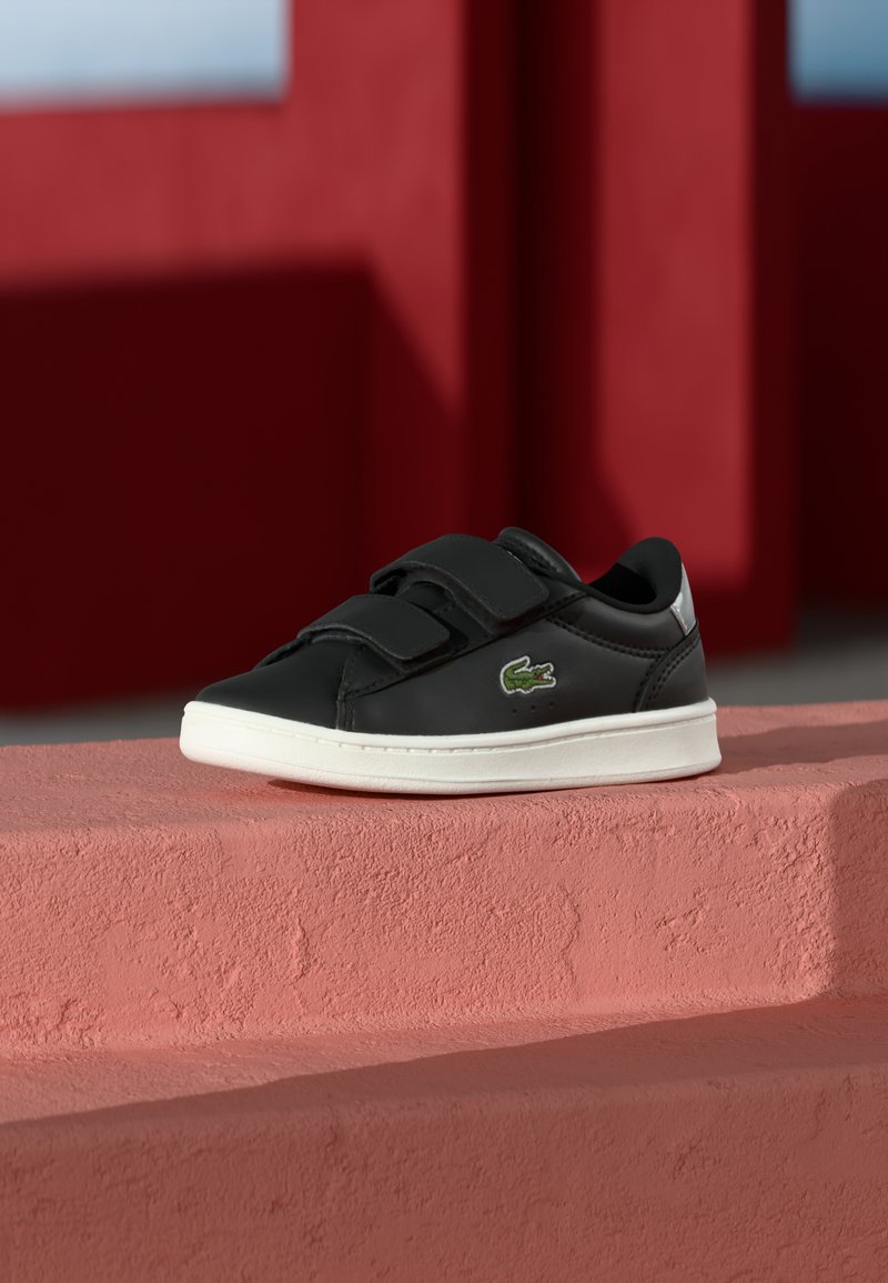 Melni ādas sporta apavi ar baltu gumijas zoli, aprīkoti ar Velcro siksnām un zaļu krokodila logo sānos. Strukturēta virsma, minimālistisks dizains.