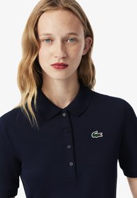 Granatowa koszulka polo z teksturowanej tkaniny z kołnierzykiem, sześcioma guzikami oraz zielonym logo krokodyla na lewej piersi. Model ma proste włosy.